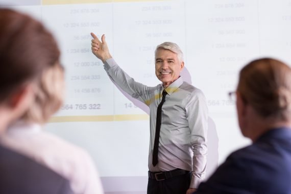 Homem de cabelos grisalhos aponta para tela enquanto ministra treinamento de vendas para uma equipe. A tela mostra alguns números.
