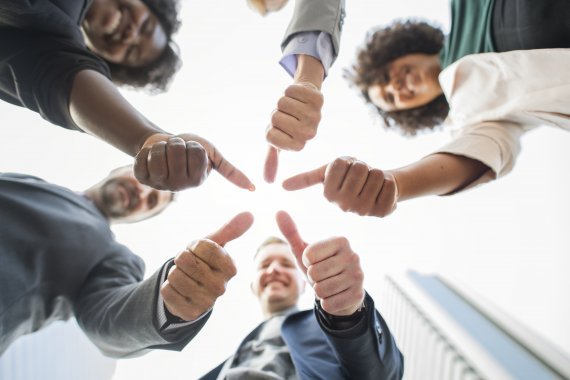 Grupo de cinco homens e mulheres sorriem e juntam os punhos com polegar para cima, em sinal de positivo. O gesto é uma demonstração de motivação no trabalho e satisfação.
