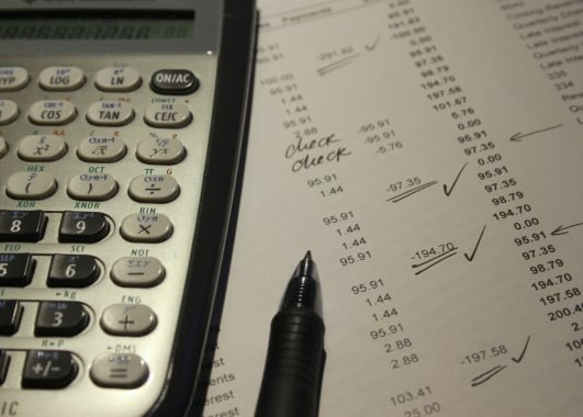 Uma calculadora e uma caneta estão sobre uma folha de papel contendo uma planilha com diferentes números. Alguns deles estão marcados com setas e confirmações. Os números representam os dados organizados em uma DRE.