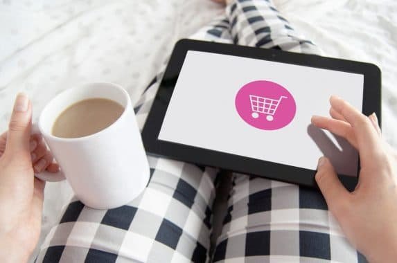 Uma mulher de calça xadrez segura uma caneca com café e um tablet no colo. A tela do tablet exibe a ilustração de um carrinho de compras, simbolizando o comportamento do consumidor digital.