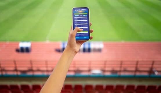 Imagem mostra mão de alto ângulo segurando smartphone com um campo de futebol à frente, simulando a importância de saber como declarar dinheiro de apostas no imposto de renda